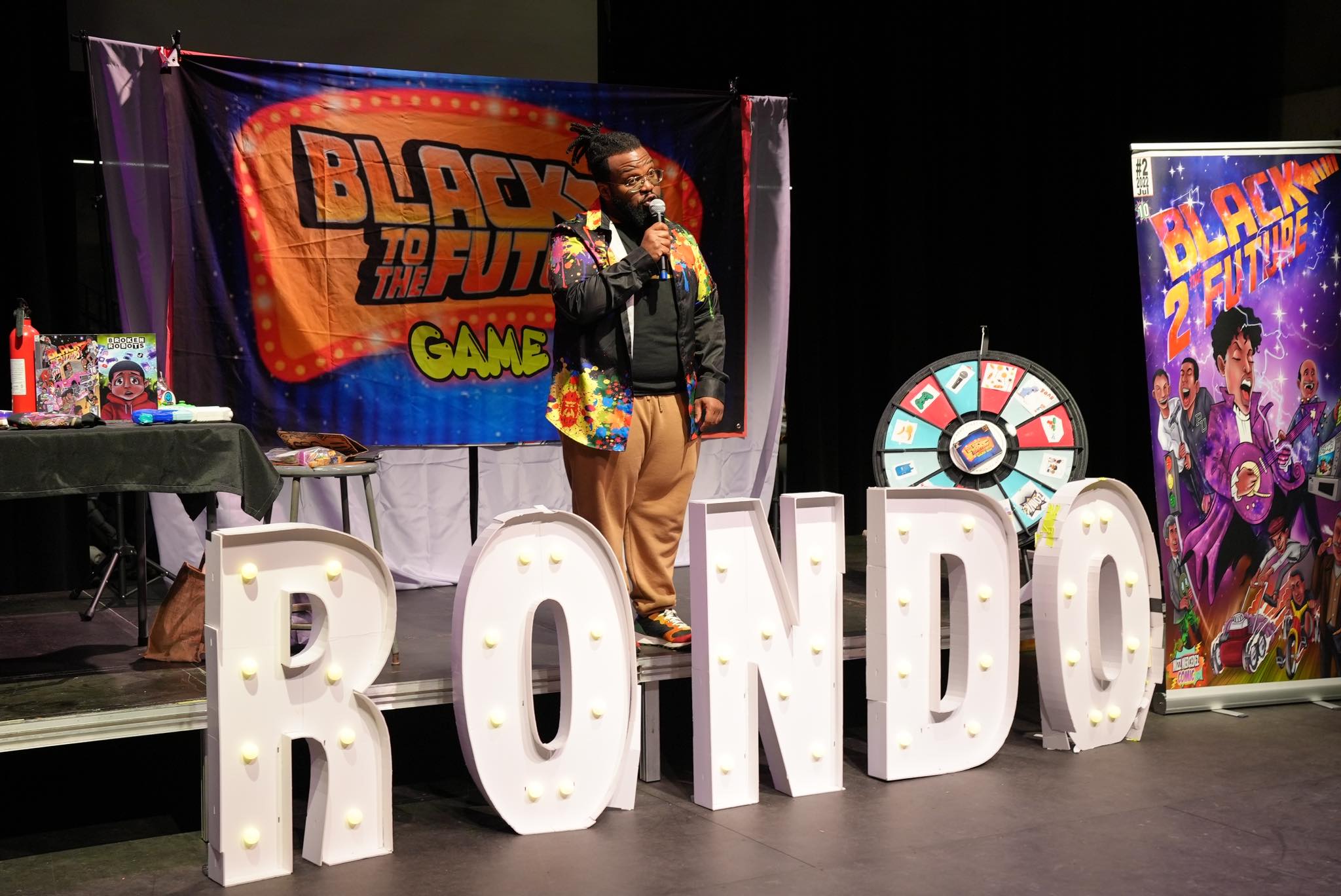 A man speaks on stage surrounded by a Rondo sign and signs for Black to the Future