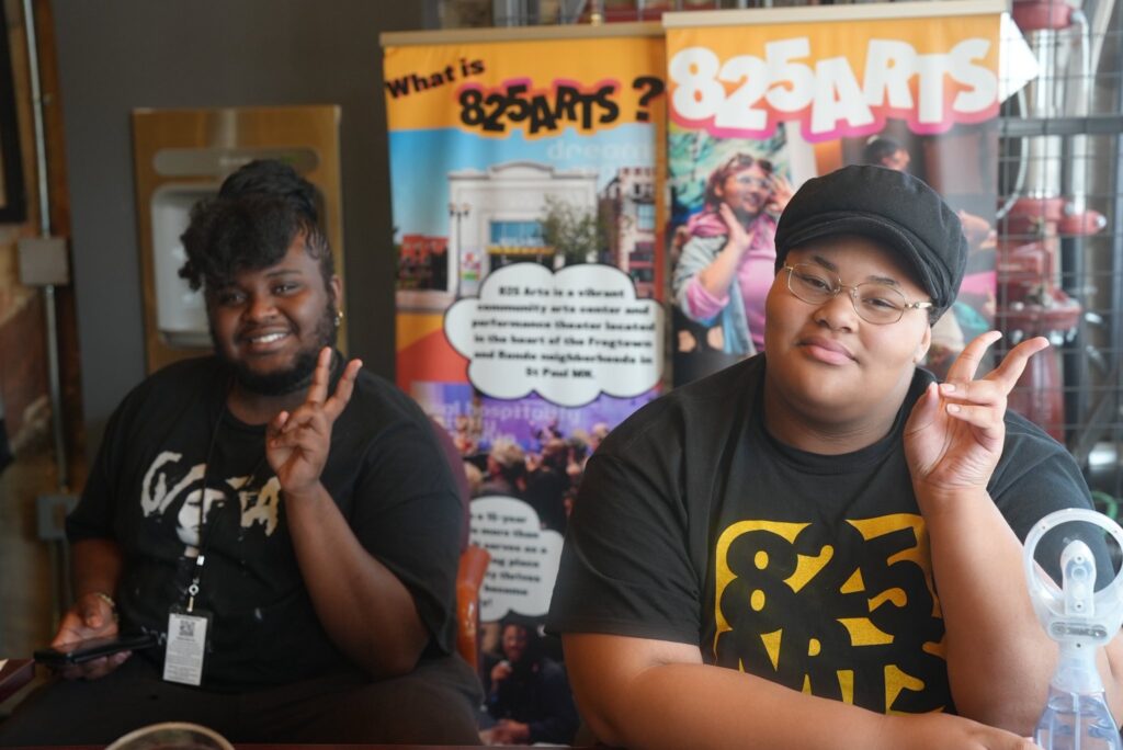 Two people hold up peace signs in front of signs for 825 Arts