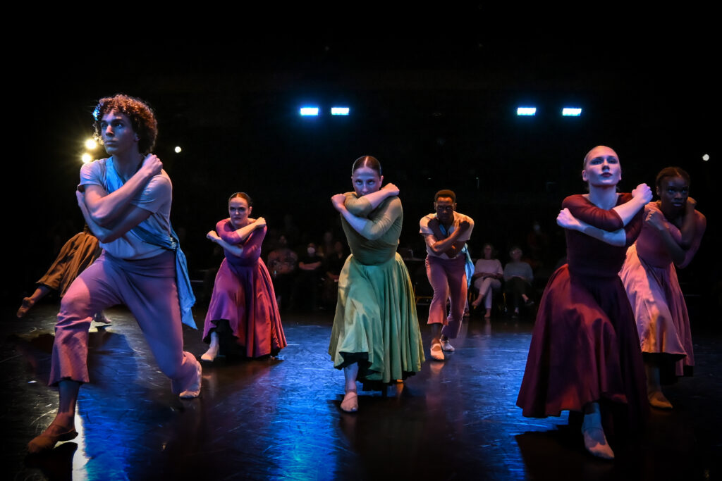 Six dancers wearing muted multicolored garments stand on a lit stage with their arms crossed over their shoulders as they stare straight ahead.