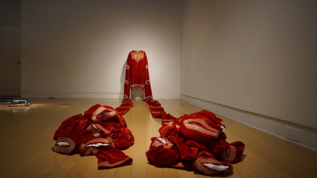 A deep red velvet garment embroidered with gold with very long sleeves that trail along the ground in two straight lines and form artful heaps of fabric at the ends.