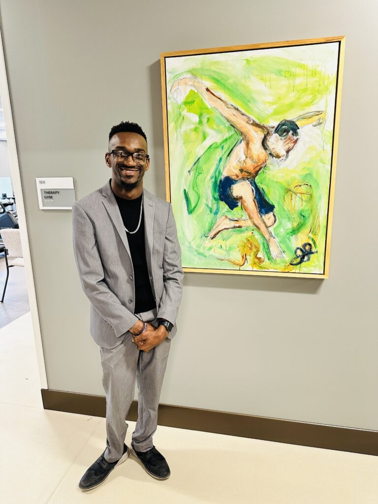 A man wearing a light gray suit and black shirt stands in front of a framed painting of a figure in motion against a bright green background. He smiles with his hands clasped in front of him.