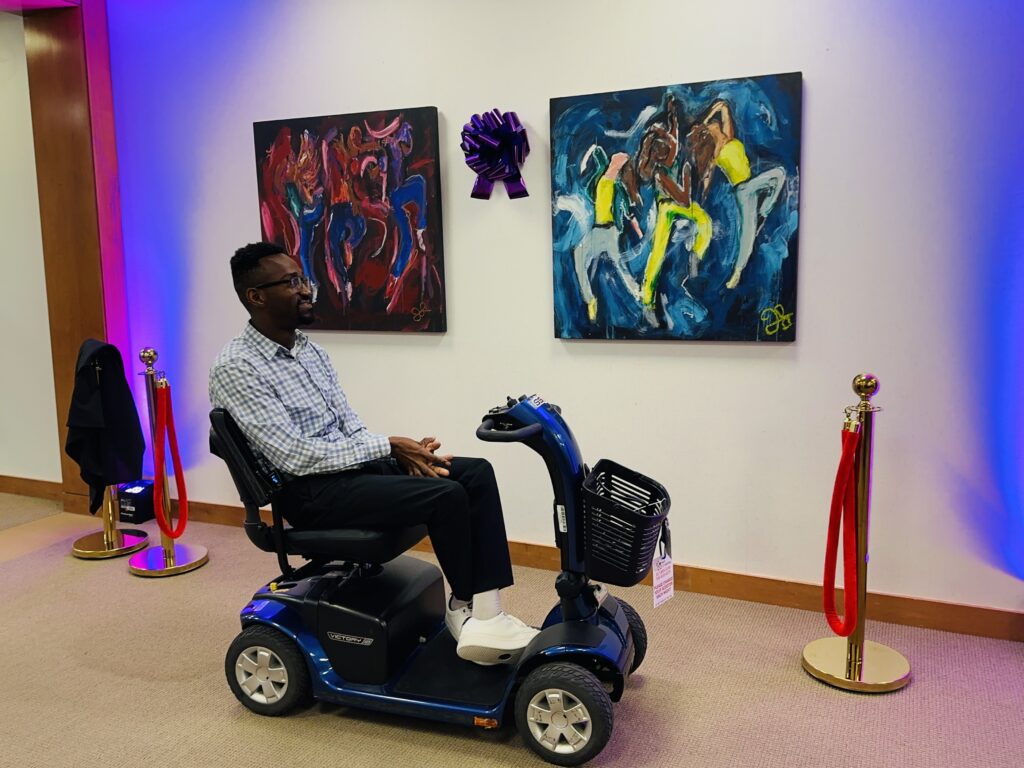 A man sits in a blue mobility scooter, smiling as he looks at two large, vibrant paintings on the gallery wall. The paintings show energetic, abstract figures in red, blue, and yellow tones.