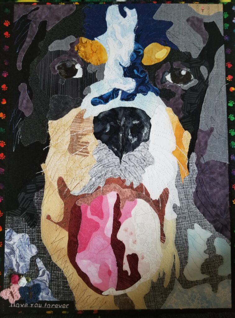 The face of a dog with long ears; black, white, and tan fur; a black nose; and a big pink tongue lolling out as it pants. In the lower left corner are stitched the words "I love you forever" below a tiny pink and a tiny blue butterfly and two tiny roses, one red and one white.