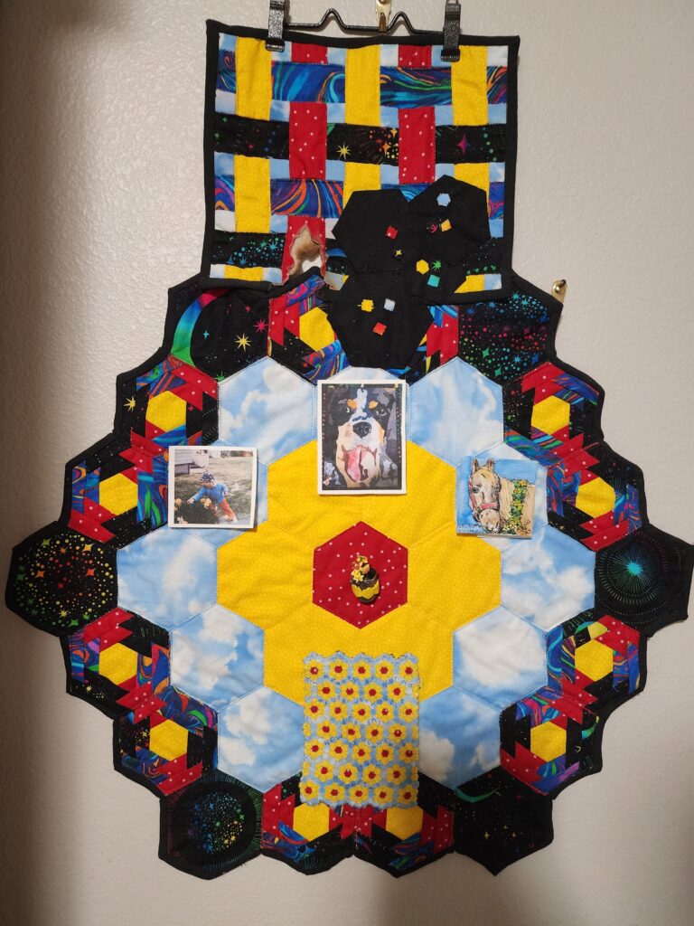 A hexagonal quilt with brightly colored and patterned fabrics around a yellow flower with a red center, adorned with pictures of a child picking daffodils, a dog with its tongue out, and a light brown horse wearing a wreath of yellow flowers. In the middle is a small pouch with tiny yellow daffodils inside.