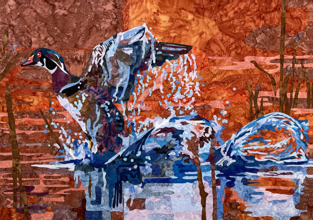 A fabric collage quilt of a duck mid flap, coming out of splashing blue water against a vibrant autumnal background