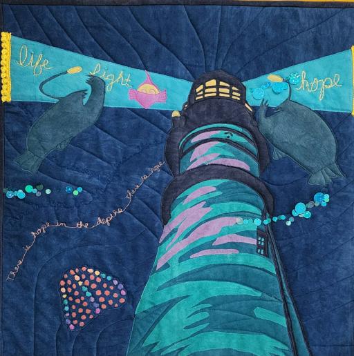 A pale cylindrical lighthouse shining two rays of light into an indigo sky with two anglerfish and other deep sea creatures floating by, along with the words "life light hope" and "there is hope in the depths there is hope" sewn in yellow thread.