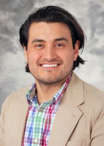 A smiling man with dark hair wearing a tan blazer over a colorful plaid shirt against a gray studio background.