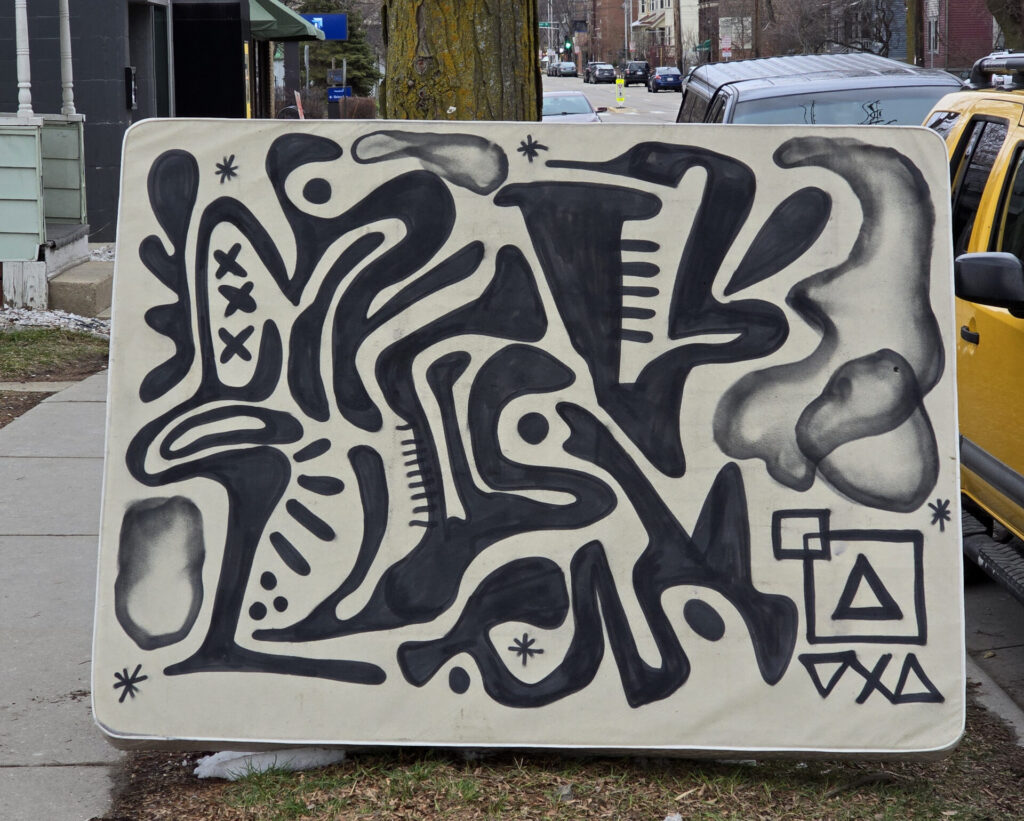 A white mattress propped up against a tree outside with black abstract art painted on it.
