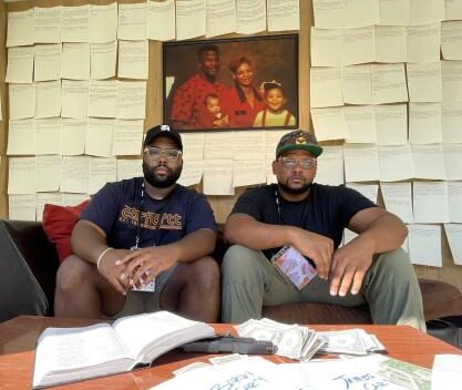 Two men with dark skin tones wearing black shirts and baseball caps sit on a couch amid writing-filled paper.