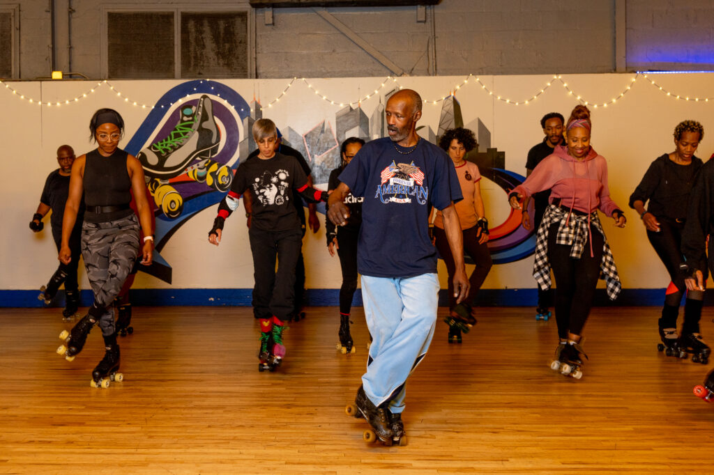 Calvin Small leads a group of skaters on a wooden rink floor, demonstrating JB roller-skating footwork as students follow behind him.