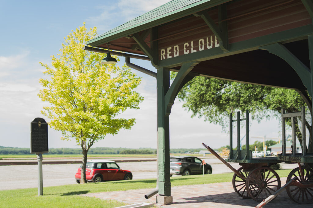 Exterior view of the Red Cloud train depot with a green roof and old baggage cart, with cars and farmland in the background.