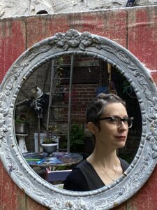 A round ornate mirror reflects the image of a femme person with short wavy hair and vintage glasses. A brick courtyard is behind her containing an array of odd objects including a skeleton puppet, a ladder, plants, and painted furniture.