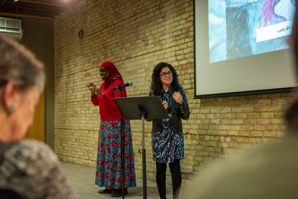 Two women stand at the front of a room with a brick wall behind them. One woman, wearing a patterned dress, black jacket, and glasses, speaks at a podium with a microphone. The other, wearing a red headscarf, red sweater, and a long blue floral skirt, stands beside her gesturing. A projected image is partially visible on the wall, and audience members are blurred in the foreground.