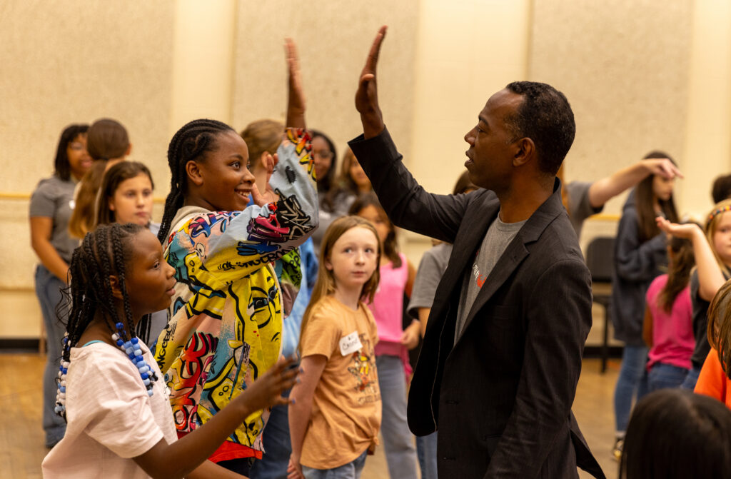 A man in a black blazer high fives a student in a brightly colored top in a room surrounded by other students.