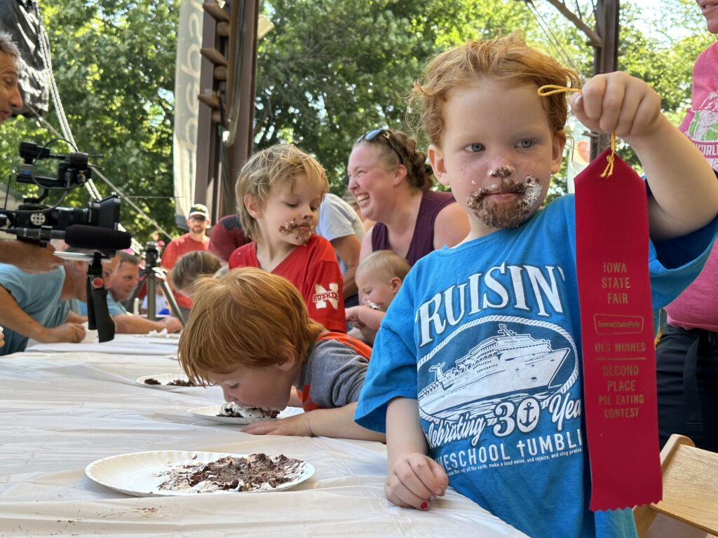 Young children at a pie-eating contest, faces smeared with pie. A child with whip cream all over their face proudly holds up a red "Second Place, pie eating contest" ribbon. Several children in the background continue eating pie, with smiling parents around.