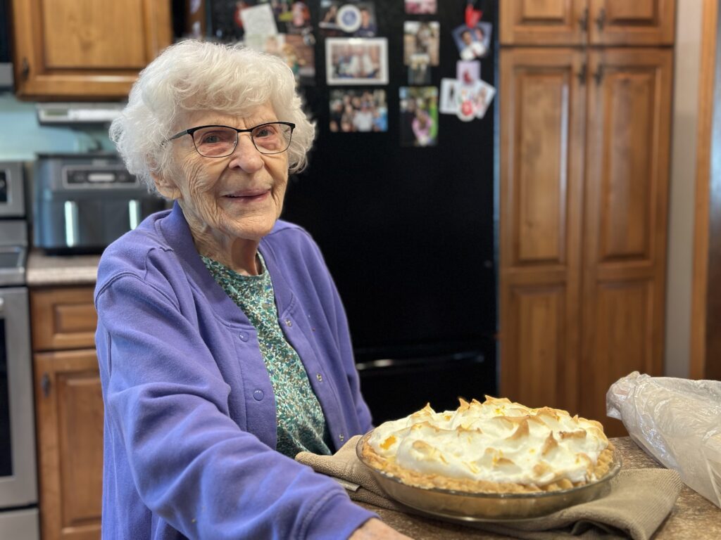 Elderly person with glasses and a purple cardigan smiles in a kitchen, proudly presenting a homemade meringue pie.