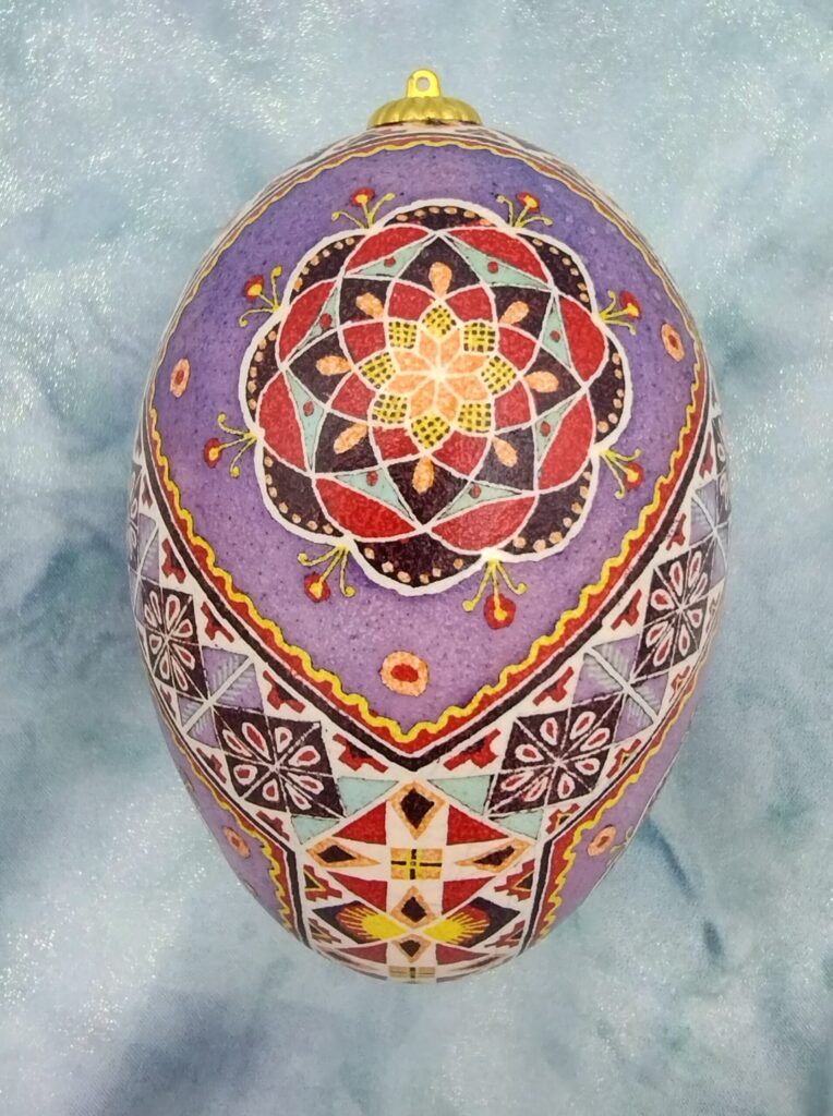 A purple egg intricately painted with geometric and floral folk patterns in red, black, yellow, and turquoise.