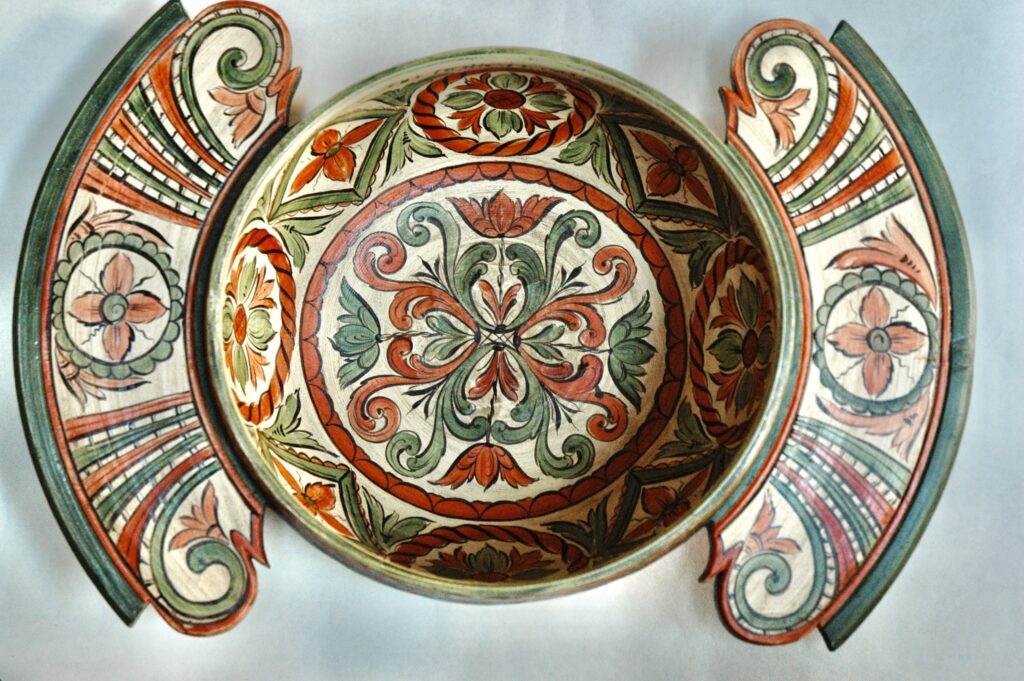 A wooden bowl with floral rosemaling designs in red, green, and gold, flanked by two matching painted panels.