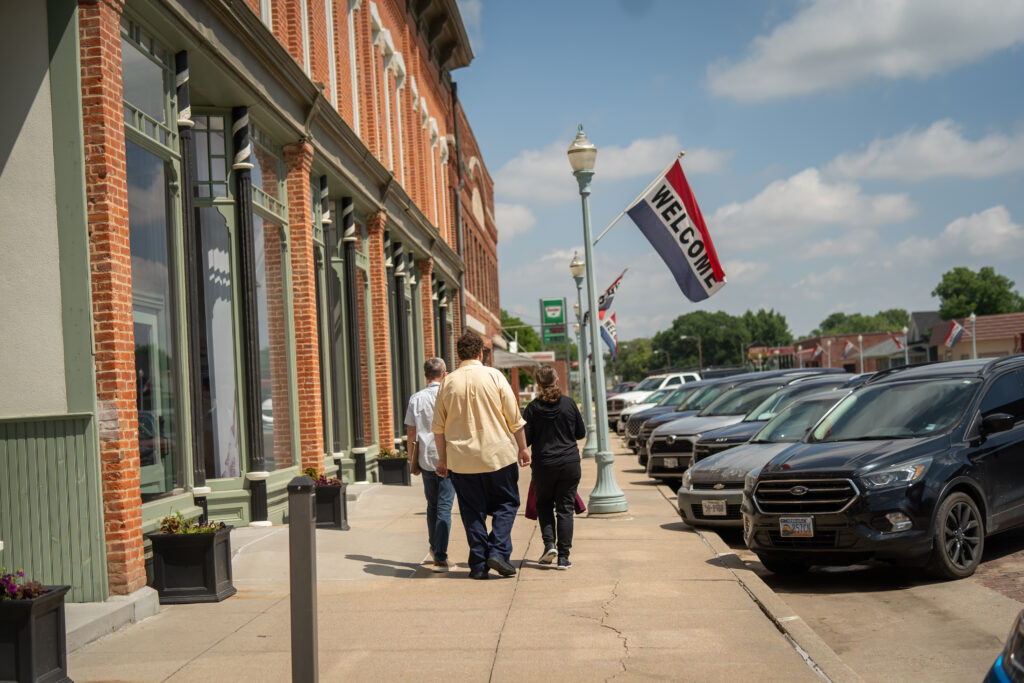 Three people walking down a small-town main street lined with brick storefronts and parked cars, with a “Welcome” flag hanging above.
