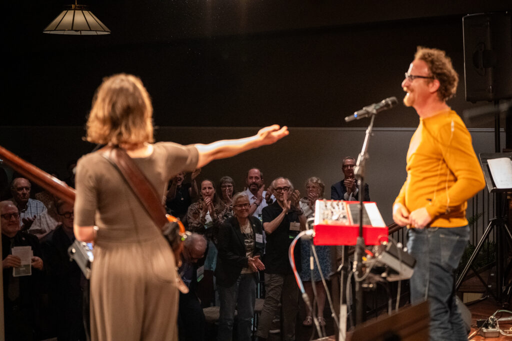 Two performers on stage in front of an applauding audience; one gestures toward the other while smiling at the microphone.