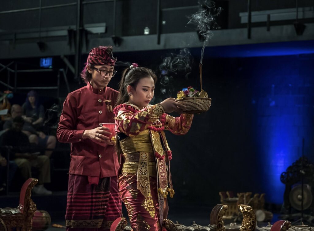 Two performers in red and gold Balinese attire prepare an offering with incense on stage.