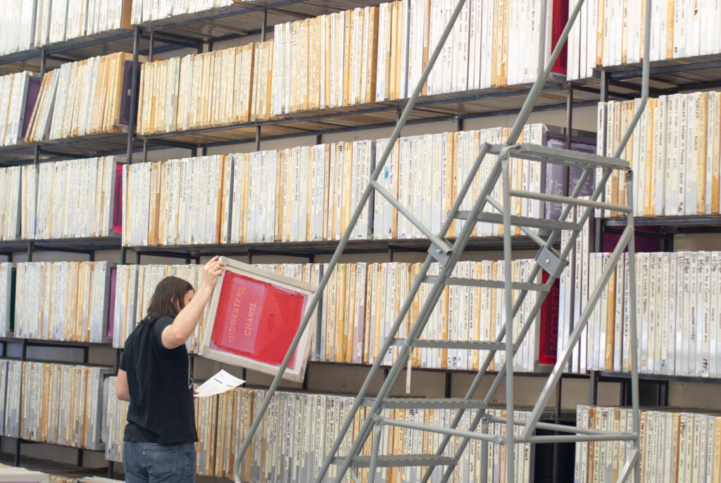 A person stands near a ladder and holds a screenprinting frame.