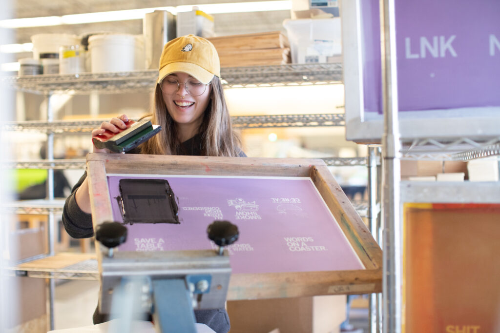 Person with long blonde hair and light skin holds up a screenprinting device and smiles.