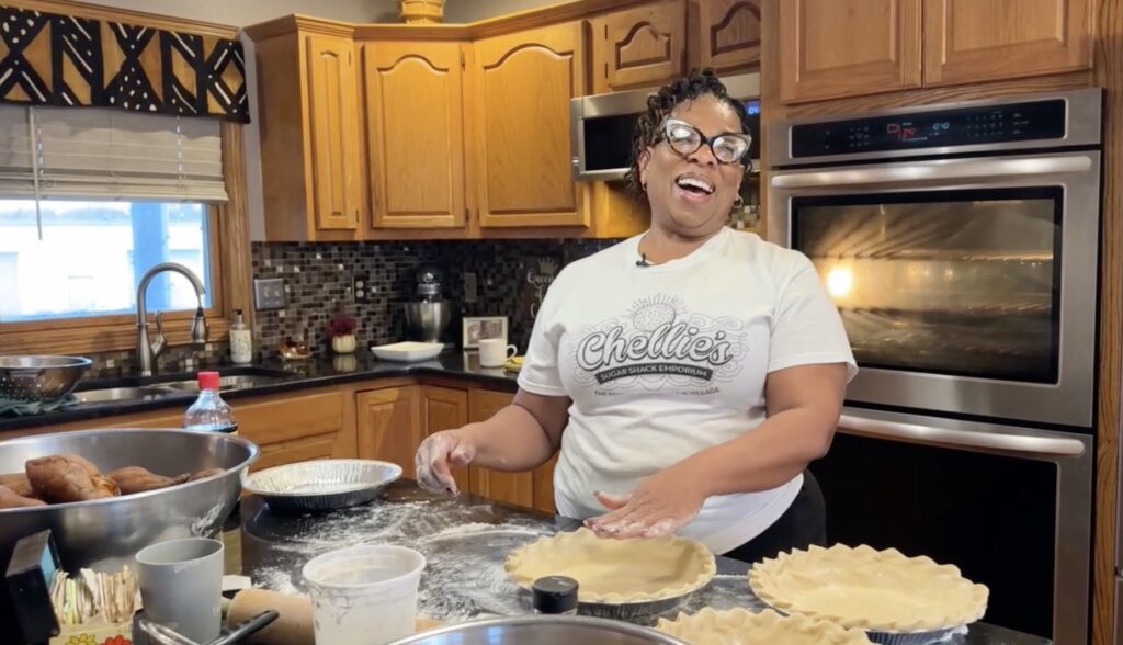 A person in a t-shirt standing in a warm kitchen, joyfully preparing pie crusts. The room is cozy, with wooden cabinets and a tiled backsplash.