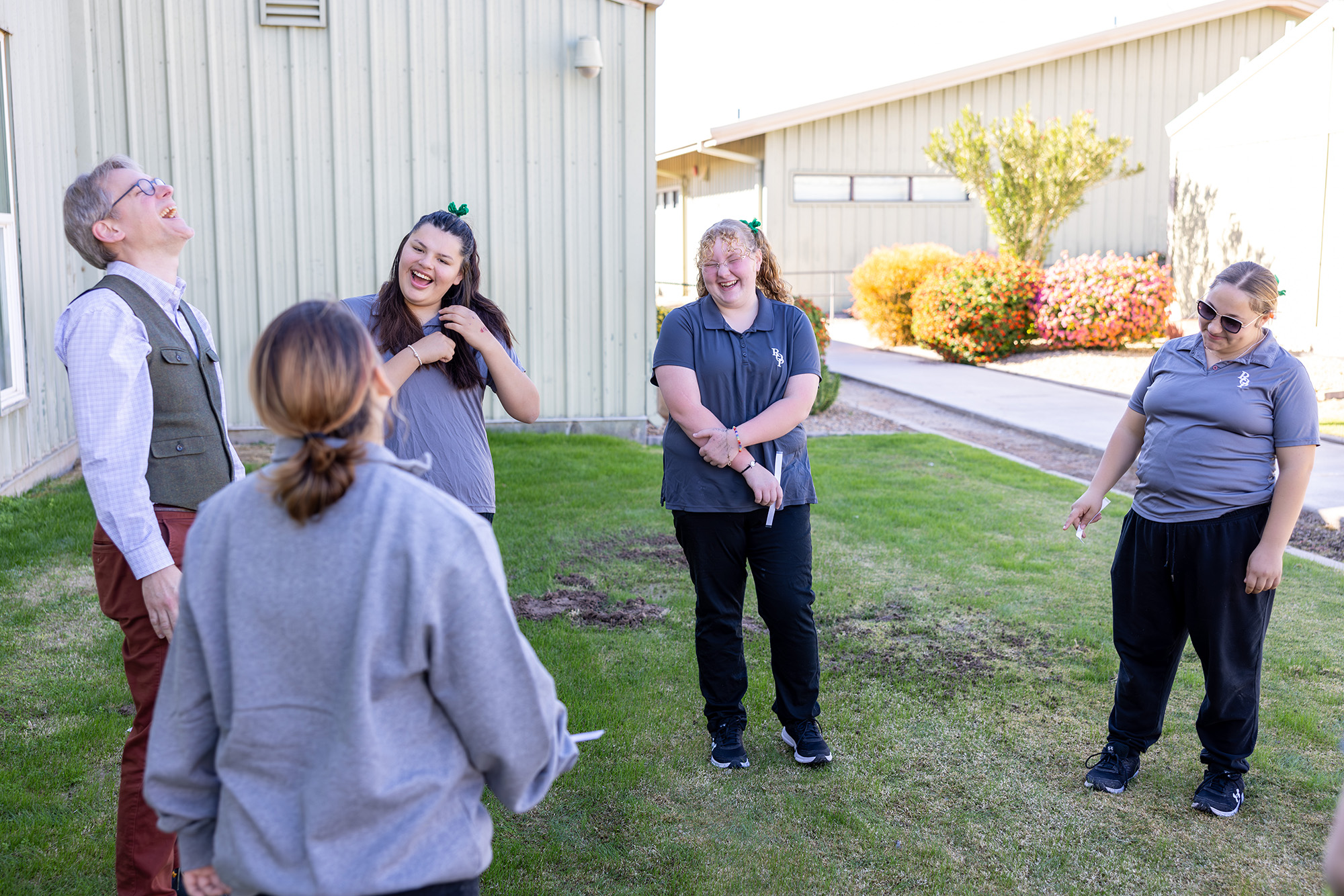 A group of five people standing outside on a lawn near a building. They are laughing and smiling.