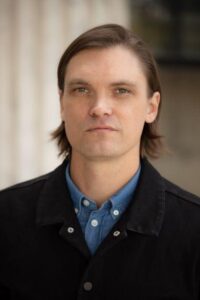 A smiling person with light skin tone and brown hair parted to one side, wearing a blue shirt and black jacket.