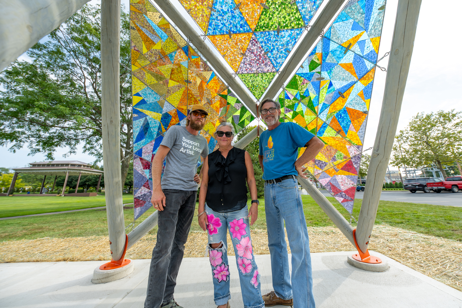 Three people with light skin tone and sunglasses standing beneath large, brightly colored glass mosaics that form an open pavilion in a public park as daylight streams through the glass behind them.