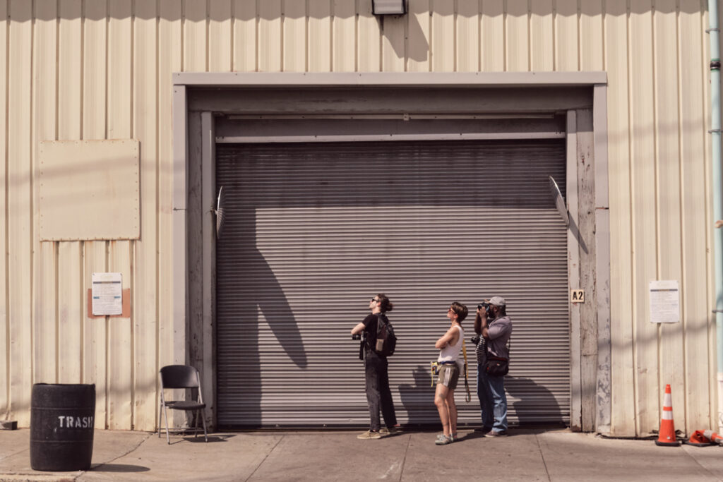Three people with cameras standing outside a closed metal garage door on a beige industrial building. One person is holding the camera up, taking photos.