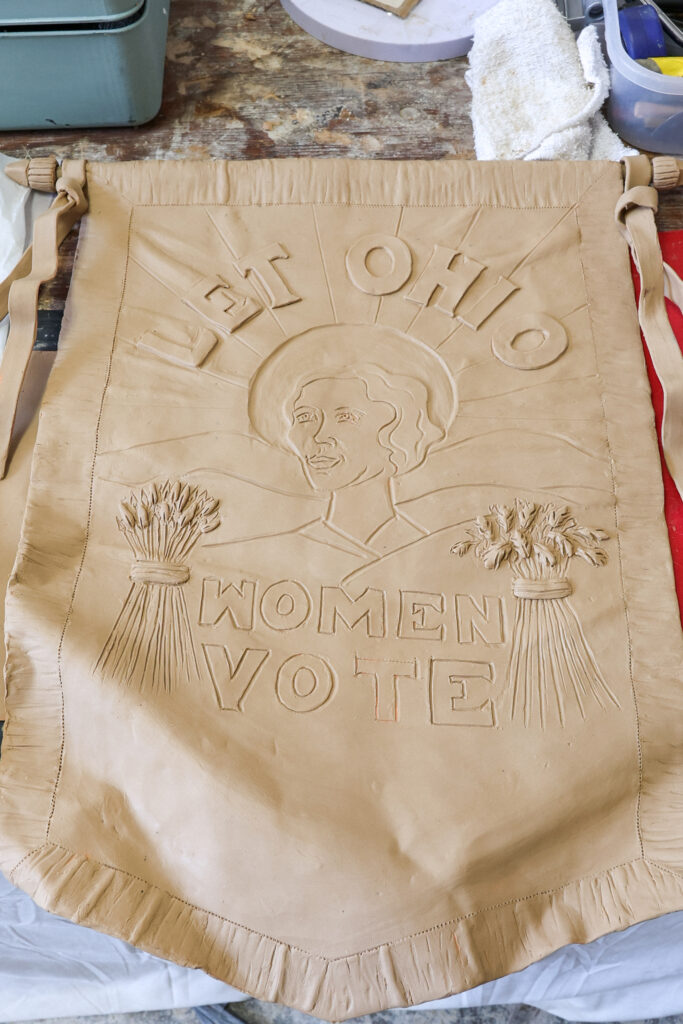 A clay relief banner reads “LET OHIO WOMEN VOTE,” featuring a portrait of a woman surrounded by sun rays and sheaves of wheat.