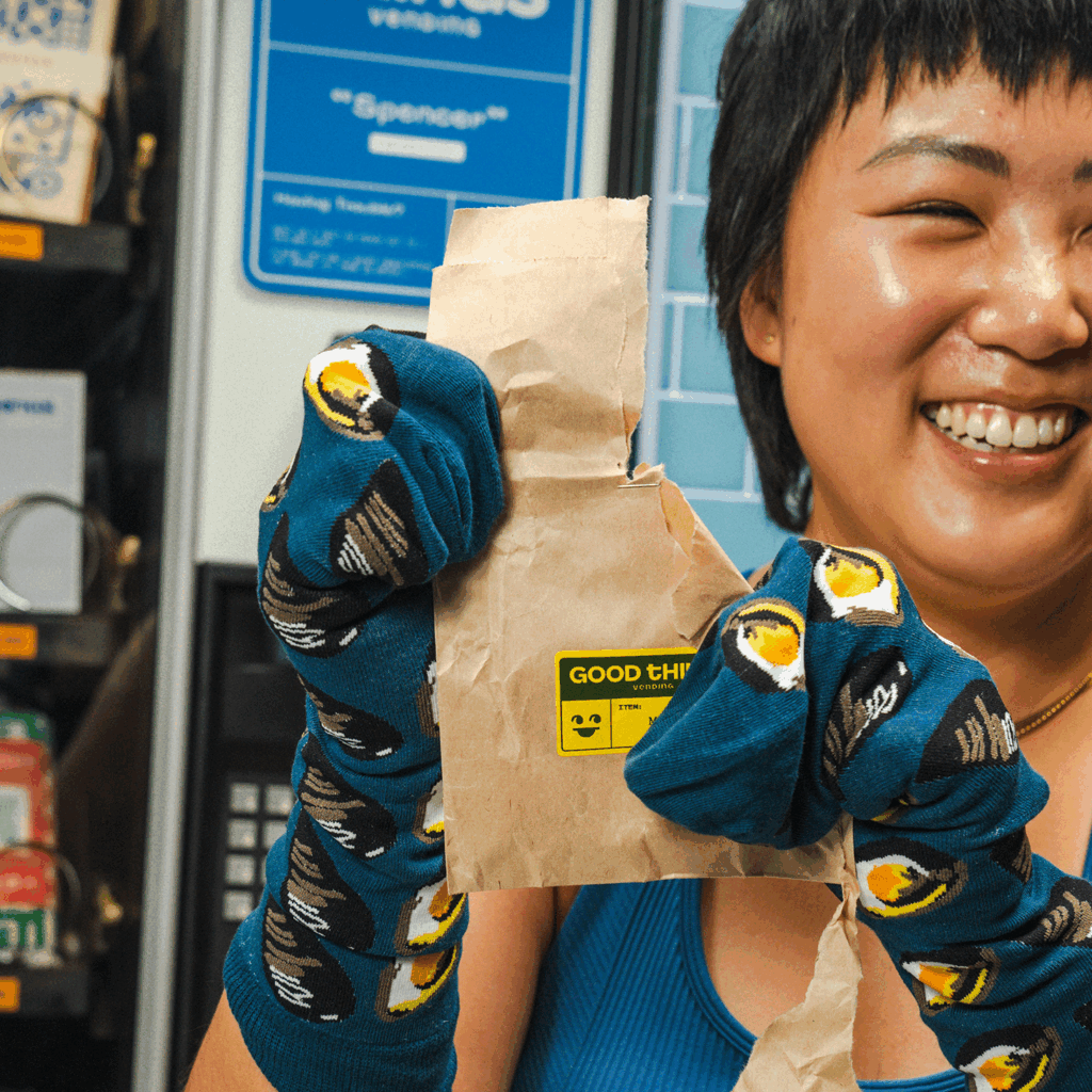 A person with medium skin tone smiles and wears socks on their hands while holding brown paper from a vending machine.