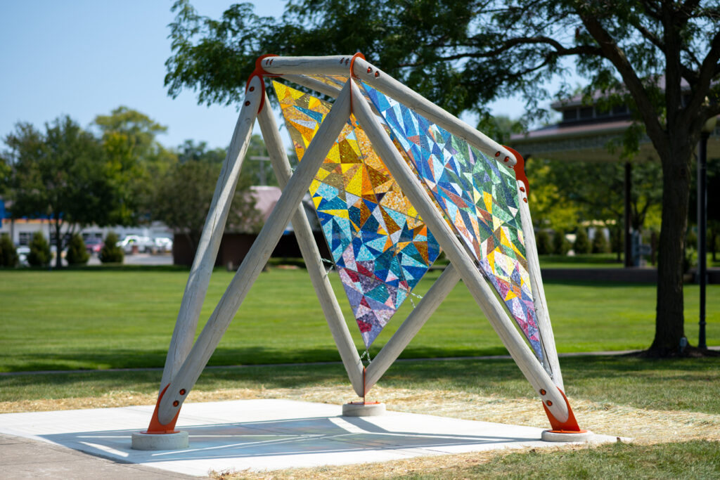 An open pavilion in a public park comprised of gray wooden beams forming large open triangles with brightly colored glass mosaics on two sides.