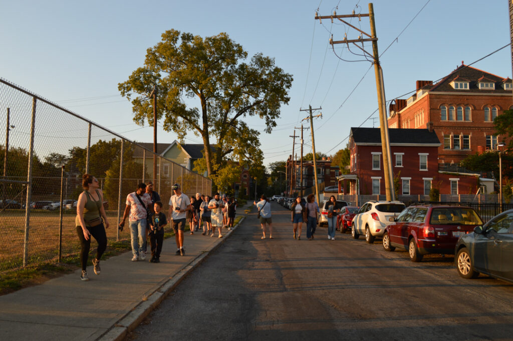 A group of people are walking down a tree-lined street in the warm glow of sunset, with cars parked on the right and a large brick building nearby.