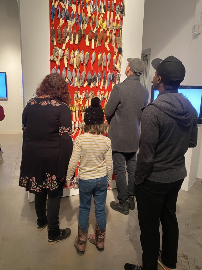 Four onlookers of varying ages looking at a large wall hanging of red cloth, with rows and rows of birds sewn from scraps of fabric tacked to the cloth with labels on each one.