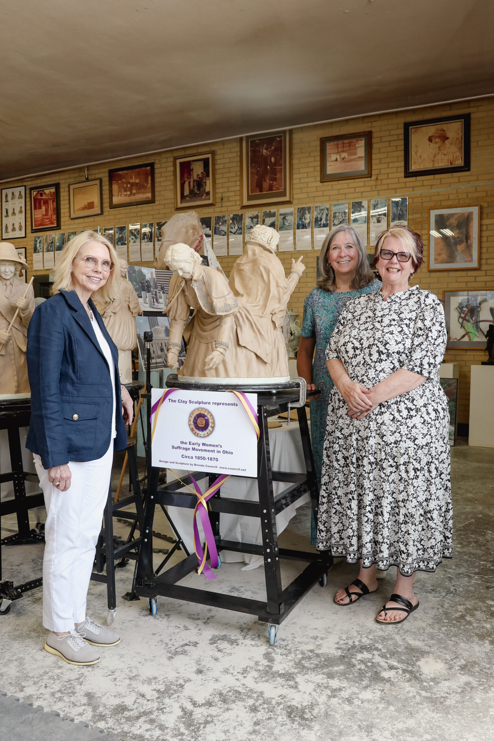 Three women stand beside clay models of suffragists in a sculpture studio. A sign reads, “The Clay Sculpture represents the Early Women’s Suffrage Movement in Ohio, Circa 1850–1870.”