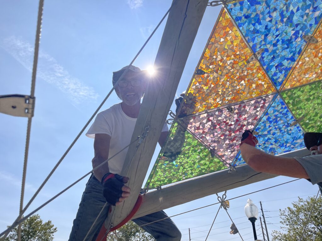 A craftsperson in a white t-shirt and jeans harnessed to a wooden support beam as they install brightly colored triangular glass panels in an open pavilion public art installation. The sun shines brightly behind them.