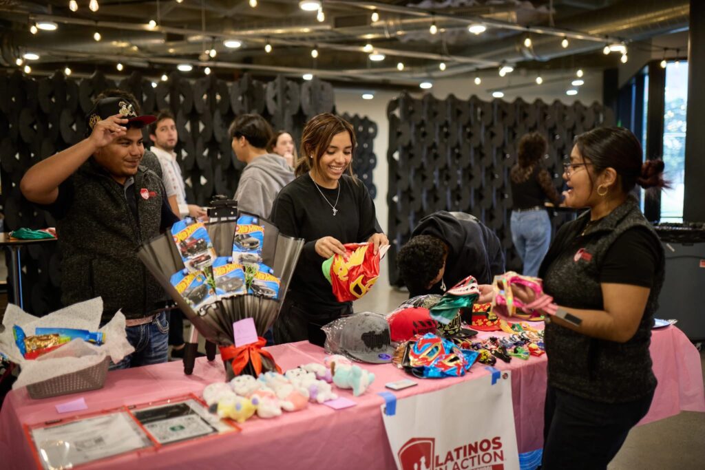 Two high school youth members of Latinos in Action standing behind a table of merchandise that includes ballcaps, luchador masks, and other colorful merchandise while a third student volunteer places more merchandise on the table.