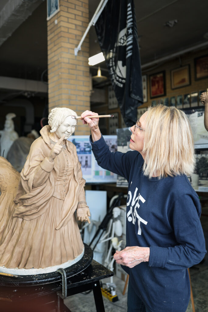 A sculptor carefully refines the facial features of a clay statue of a suffragist woman, adding detail with a small metal tool.
