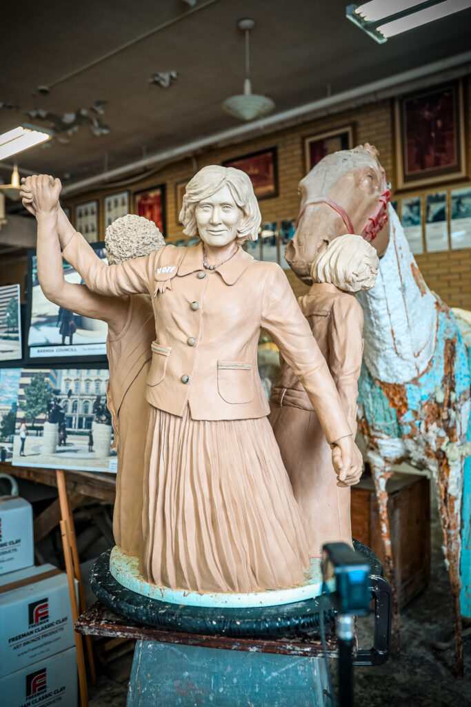A nearly complete clay sculpture of a woman in mid-century clothing holds hands with others in a circle. A “VOTE” button is visible on her jacket.