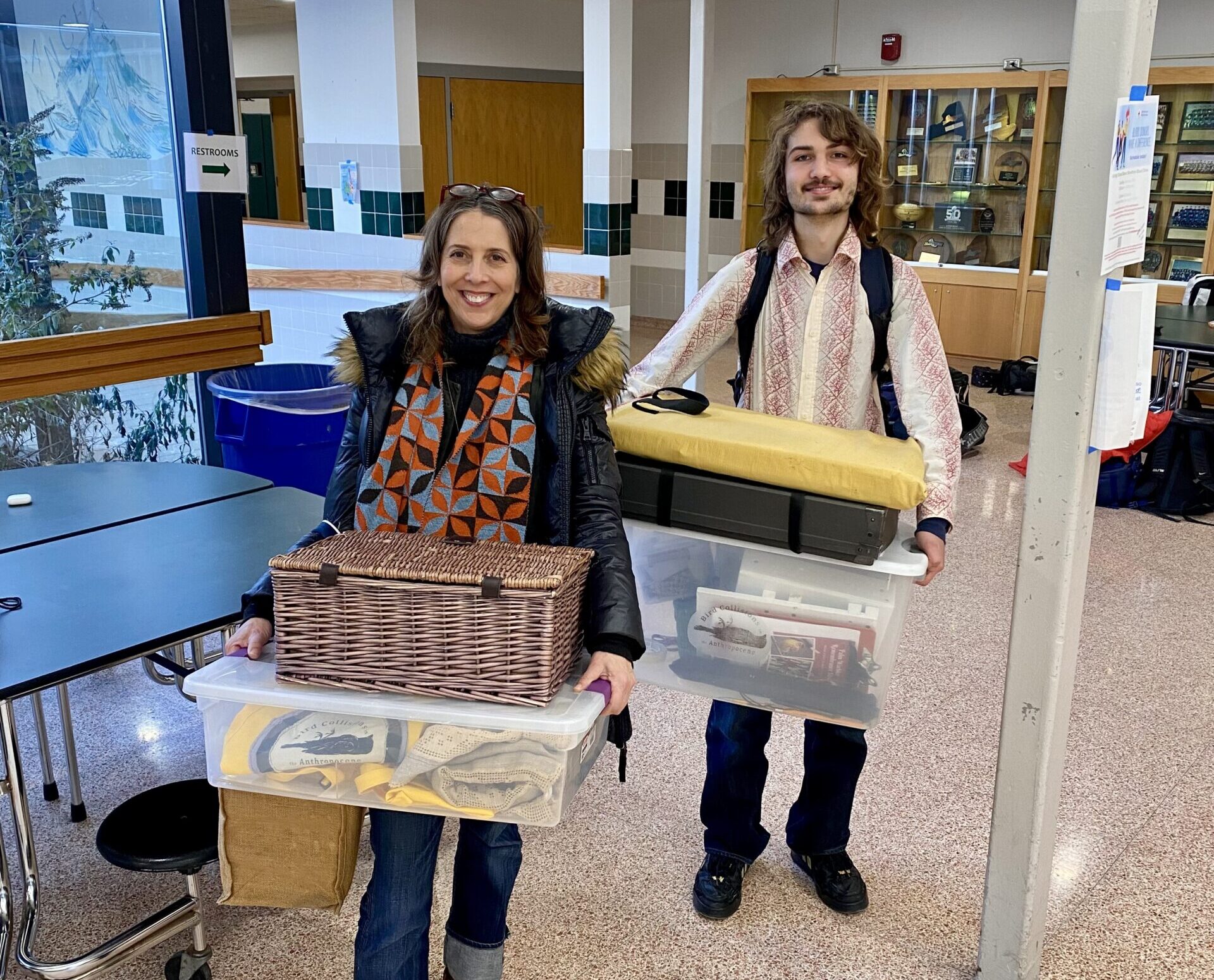 A woman with medium-length brown hair and a young man with long brown hair and a beard smiling as they carry bins of sewing supplies.