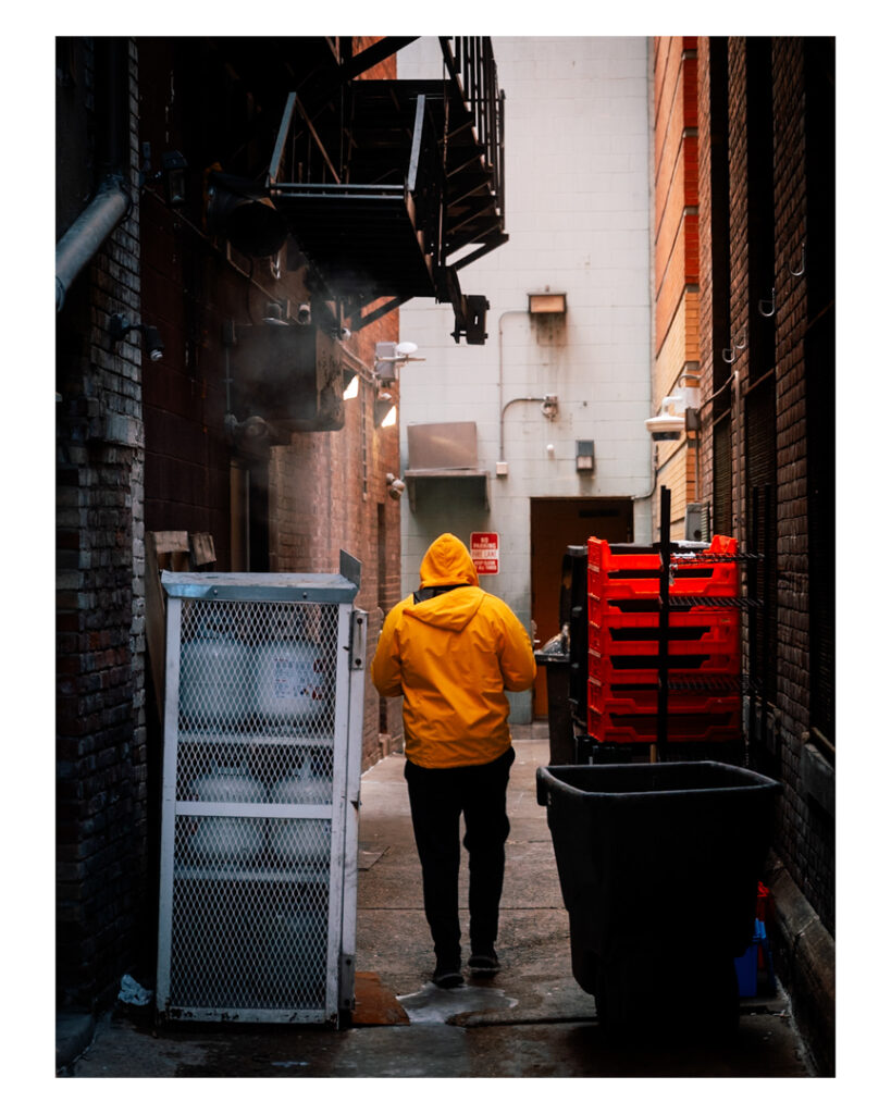 A person wearing a bright yellow hooded jacket and dark pants walking down a narrow urban alley lined with brick walls, metal staircases, and trash bins on a cloudy day