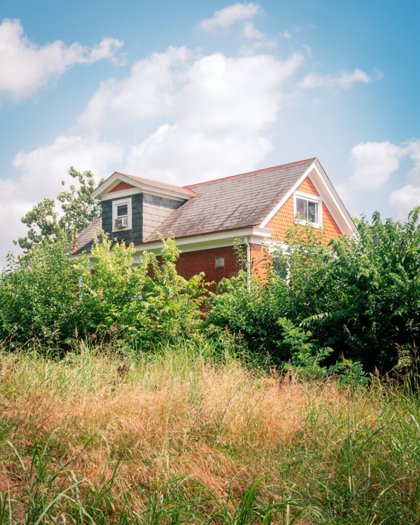 A quaint house with red siding and a gray roof is partially obscured by lush greenery. It rests under a bright, blue sky with scattered clouds.
