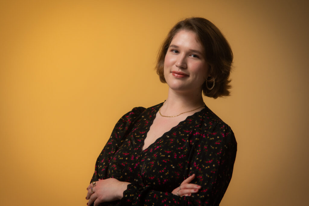 Headshot of a smiling person with light skin tone and chin-length light brown hair, wearing a floral print dress, gold hoop earrings, and a gold chain necklace.