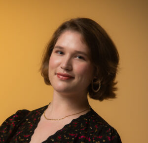 Headshot of a smiling person with light skin tone and chin-length light brown hair, wearing a floral print dress, gold hoop earrings, and a gold chain necklace.