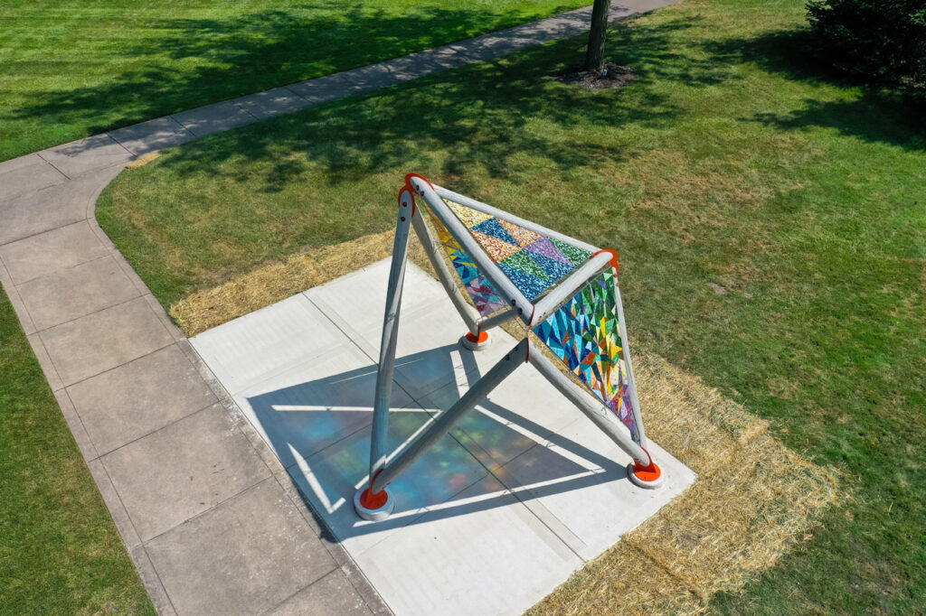 An aerial view of an open pavilion constructed of wooden beams supporting brightly colored mosaics of glass triangular panels in a public park.