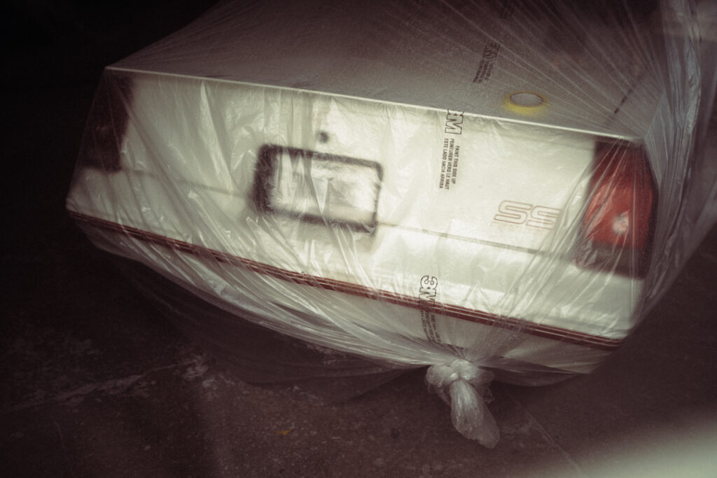 A white car is partially wrapped in clear plastic, parked in a dimly lit area.