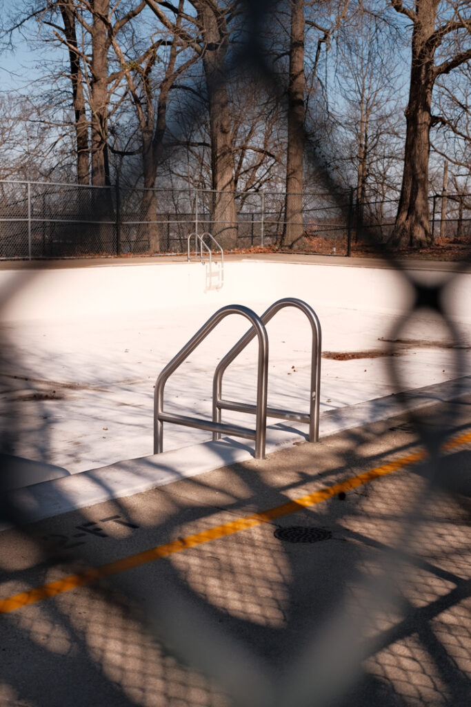 Empty swimming pool with dry leaves, surrounded by bare trees and a chain-link fence. Sunlight casts shadows of the fence and trees.